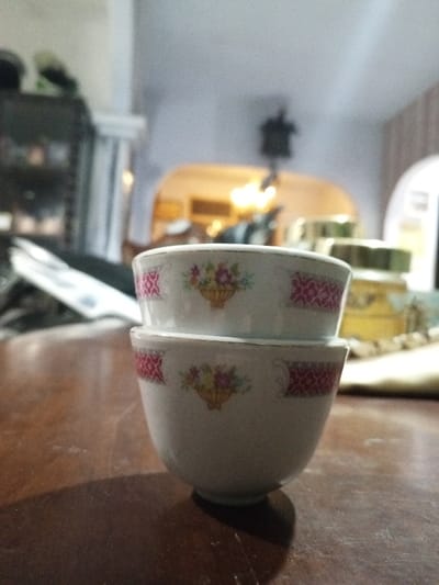 The image displays two small, white, ceramic bowls stacked centrally on a dark brown wooden table, in an indoor setting in Longmiang, Laos. Each bowl features an identical repeating pattern of a central floral bouquet in a yellow basket, flanked by two magenta geometric lattice designs. A thin gold-colored line outlines the rim of both bowls. The wooden table surface shows faint scuffs or dust. The background is significantly blurred, revealing an arched doorway leading to an adjacent room illuminated by warm, yellow light. To the right of the stacked bowls, out-of-focus objects include two cylindrical containers with golden lids, one appearing to hold a yellow substance, placed near a light-colored fabric item. To the left, a dark, indistinct furniture piece and a vertical light-colored structure are partially visible. A dark object is vaguely suspended from the ceiling above the arched opening. No people are present, and no actions or interactions are depicted.