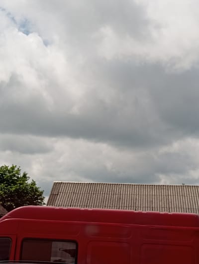 The image shows a portion of a red van parked outdoors under a cloudy sky. To the right of the van, a roof made of corrugated material is visible, with a hint of green foliage from a tree on the left side of the frame. The scene is set in Hlynytsi, Ukraine.