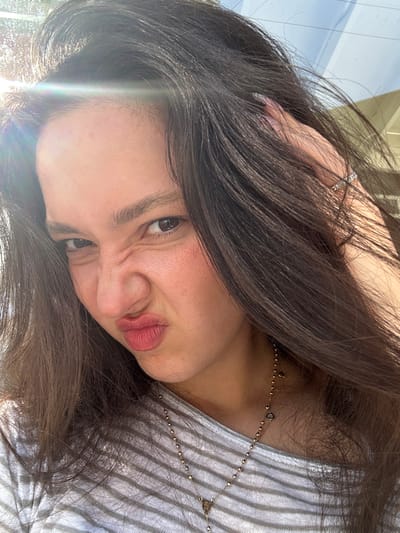 This image captures a close-up portrait of a female subject in Glendale, United States. The individual has long, dark brown hair and a fair to medium skin tone. Her face displays an exaggerated expression, characterized by a crinkled nose, slightly furrowed eyebrows, and pursed lips, commonly known as a "duck face." Her dark brown eyes are directed at the viewer. Her right hand is visible in the upper right, fingers gently touching her hair. On her right ring finger, she wears a silver-colored ring, possibly an eternity band with small stones. She is dressed in a grey and white striped top and a gold-colored necklace with small spherical beads and at least two heart-shaped charms. Bright sunlight, producing a prominent lens flare in the upper left, illuminates the scene, indicating an outdoor setting. A light-colored, angular structure is faintly visible in the background, suggesting an architectural element like an awning or building.