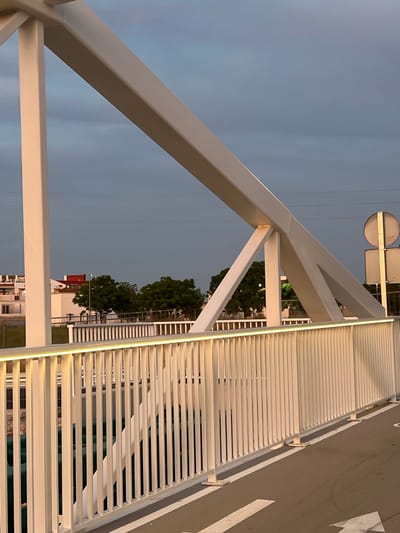 This image depicts a modern, white-painted metal truss bridge located in Alcàsser, Spain, during what appears to be twilight or dusk. The bridge's superstructure features prominent, rectangular cross-section beams arranged in a truss configuration, with an inclined main beam supported by vertical and diagonal struts. A white metal railing runs along the visible side of the bridge, composed of numerous vertical balusters. A warm, yellow LED strip light is integrated along the top edge of this railing, illuminating the structure. Below the railing, a paved path or road surface is visible, marked with white directional arrows, one partially visible pointing towards the lower right.

Through the gaps in the bridge's structure and railing, a body of water, possibly a canal or river, can be seen with a dark greenish hue. Beyond the bridge on the left, white residential buildings with terracotta-colored roofs are visible against the horizon. Clumps of green trees are interspersed between the buildings and the waterway. In the upper background, thin horizontal lines, indicative of overhead power lines, traverse the scene. On the right side of the bridge, the back of a circular road sign and a rectangular sign, both mounted on a metal pole, are partially visible. The sky overhead is a uniform, muted blue-gray, suggesting overcast conditions or the time of day. No people or animals are present, and no specific actions or interactions are depicted.