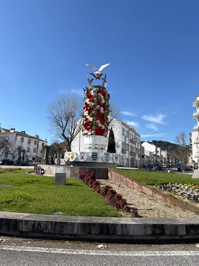A tall, ornate monument stands at the center of a landscaped traffic roundabout in Tomar, Portugal, under a clear blue sky. The monument features a white cylindrical base, encircled by a band of relief work and a series of heraldic shields. Above this, the main column is densely decorated with red and white sculpted floral elements, appearing to be poppies, interspersed with numerous sculpted human faces. The monument is crowned by a golden, open-style crown, from which a white dove with outstretched wings appears to ascend.

The roundabout itself is covered in green grass, with sections of gravel and parallel rows of small, reddish-leafed plants. A low concrete curb defines the perimeter of the central island.

In the background to the left, several multi-story buildings with white facades and traditional architecture line a street, where multiple vehicles are parked. Bare deciduous trees are visible, along with what appears to be a large, weathered stone archway structure. To the right, additional multi-story white buildings extend along a street with active vehicular traffic. A distant hill, densely covered with trees, forms the horizon line. The sky is bright blue with scattered thin, white cirrus clouds, indicating clear weather. No individuals are visible in the immediate scene.