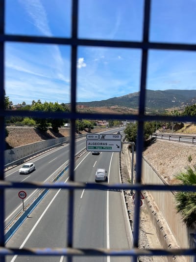 The image captures a view of a highway in Algeciras, Spain, as seen through a metal grate. The perspective is from above, looking down onto the road. The sky is a clear, bright blue with some wispy clouds and a contrail. It appears to be a sunny day.

The highway has multiple lanes with white lane markings and a blue striped divider. Several vehicles are visible on the road, including a white van and a white car. A speed limit sign indicating "60" is prominently displayed. Road signs provide directional information, with one indicating "ALGESIRAS oeste" and "400 m" followed by "avda. Agua Marina" and an arrow pointing to the right. Another sign has the number "1120" and an arrow.

The surrounding landscape features a hillside covered in vegetation and trees, with some buildings visible in the distance. The embankment along the highway is reinforced with concrete and has a rocky texture. A palm tree is visible in the upper right corner. The overall scene depicts a typical day on a Spanish highway.