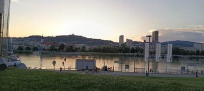 This is a panoramic, eye-level, wide-angle photograph of the Danube River waterfront in Linz, Austria, during sunset. The sky is a soft gradient of pale yellow and orange, with some scattered clouds. On the left side of the frame, a modern glass building is partially visible. The river occupies the middle ground, reflecting the warm light of the sunset.
Across the river, the Linz cityscape stretches along the riverbank, featuring a mix of historic buildings, including a church with a spire, and modern high-rise structures. A prominent hill rises behind the city, topped with a structure that resembles a monastery or church. The river is dotted with several boats, including a white boat with the name "SAVOR CH BASIL" on its side. A white trailer or small building is situated on the waterfront promenade.
On the right side of the promenade, several tall, white banners with blue logos and text reading "DONAUSCHIFFFAHRT WURM & NOE" are displayed. A street lamp is positioned next to these banners. A metal fence separates the promenade from the river.
In the foreground, a grassy area with scattered small white flowers slopes down towards the river. A few people are visible on the promenade. On the left, a child walks with an adult. Further to the right, two people are on bicycles near the trailer. Another person is walking further down the promenade on the right side. Bicycles are parked near the promenade, and a few benches are visible along the waterfront. The overall atmosphere is calm and serene, indicative of a late summer evening.