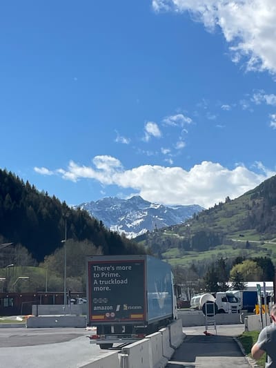 The image shows a landscape with mountains in the background partially covered in snow. In the foreground is an Amazon truck, with an advertisement on its side that says "There's more to Prime. A truckload more." The truck is located on the side of a road, with other vehicles and infrastructure present. A partly cloudy sky dominates the upper left portion of the frame. Based on visual cues, the location appears to be near the town of Freienfeld - Campo di Trens, Italy, as indicated by the landscape features.