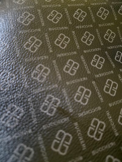 The image shows a close-up, angled view of a textured, dark green surface embossed with a repeating pattern. The pattern consists of the word "BUCCHERI" in white lettering, oriented in various directions, interspersed with a stylized white logo that resembles an interlocking "B" or an abstract symbol. The surface appears to be leather or a similar material, with a visible grain or texture. The lighting creates shadows that highlight the embossed elements and the texture of the material. No specific location within Bireuenmeunasahblang, Indonesia, can be determined from the visual information presented.