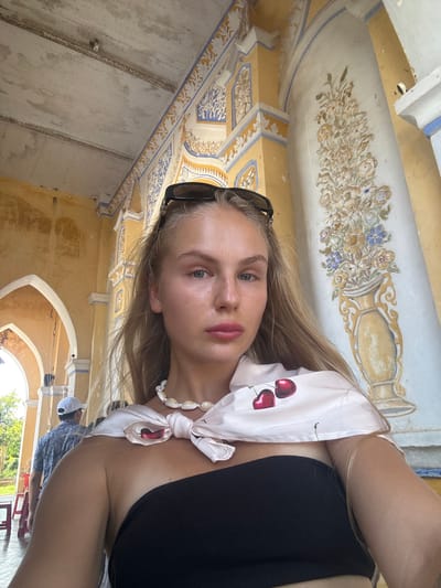 A young woman with long blonde hair is captured in a selfie, her head tilted towards the camera. She is wearing a black top and a light pink scarf tied around her neck, adorned with a cherry print. Sunglasses are perched on her head. Her expression is relaxed and serene.

The background reveals an ornately decorated interior, likely a historical building. The ceiling and walls are adorned with intricate paintings and patterns in yellow, white, blue, and gold hues, featuring floral motifs and what appear to be classical urns. The architecture suggests a traditional or historic Vietnamese setting, possibly a temple or an old villa, consistent with the provided location context of Hội Tín, Vietnam.

Sunlight streams in from an unseen source, illuminating the scene and creating soft highlights on the woman's skin. The overall impression is one of a peaceful moment captured during a visit to a culturally rich location. There is no visible text in the image.