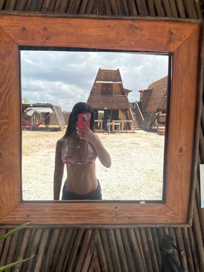 A wooden-framed mirror is mounted on a wall of woven wooden sticks. The mirror is reflecting a woman in a pink bikini, taking a selfie in front of a two-story wooden building with a thatched roof. The woman is standing on a sandy surface in front of the building. The image was taken at the coordinates 12.201, -72.148, which is in the city of Cabo de La Vela, Colombia.