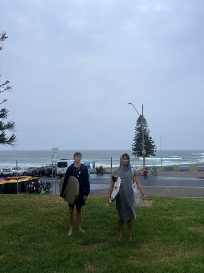 This image depicts two young adult males standing barefoot on a grassy incline at Bondi Beach, Australia, under a uniformly overcast, light grey sky. The male on the left, with short light-brown hair, wears a dark hooded long-sleeved top and dark shorts, holding a light-colored foam surfboard under his right arm and wearing white earbuds. The male on the right, with dark hair partially covered by a hood, wears a grey hooded changing poncho and carries a white surfboard with black fins under his left arm, along with a light-colored tote bag over his right shoulder; his right hand is slightly extended, palm facing forward.

Behind the subjects, a paved walkway and road separate the foreground from a coastal environment. To the left, a parking area contains various vehicles, including multiple cars and a white van, with bicycles parked near green waste bins. A white sign mounted overhead reads "BONDI BEACH PARKING AREA." To the right, a tall, slender Norfolk Island Pine is visible, featuring a street light fixture, adjacent to additional paved ground with scattered colorful concrete street furniture. In the background, the grey-blue ocean is active with numerous breaking waves, leading to a visible strip of sandy beach where distant figures are sparsely distributed. A portion of a dark green coniferous tree is visible on the far left edge of the frame.