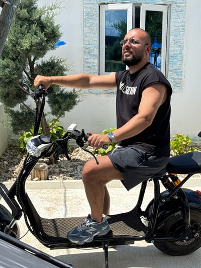 In Baliovtsi, Bulgaria, a man with a beard and glasses, wearing a black t-shirt with the word "VALUATION" in white lettering and dark gray shorts, sits on a black electric scooter. He has a watch on his left wrist and a small star tattoo on his right bicep. The scooter has a digital display on the handlebars and a reflective headlight. The man is holding the handlebars, with his left hand on the brake lever. He is looking to his right. The background features a white building with blue trim around the windows, a small evergreen tree, and a ground covered in small pebbles. The scene appears to be outdoors on a sunny day.