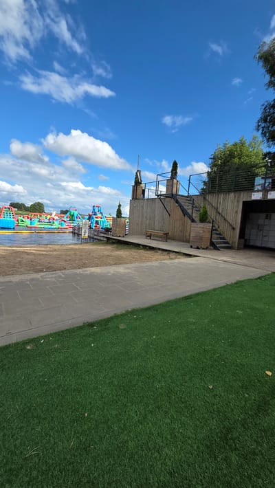 An outdoor scene features a large inflatable water park on a lake in the background, with multiple colorful slides and obstacles. In the foreground, there is a sandy area transitioning to a paved walkway and then an area of artificial turf. To the right, a wooden structure with stairs leads up to a platform. There is a bench on the wooden platform. The sky is blue with scattered white clouds. The location is identified as being in Jaunmārupe, Latvia.