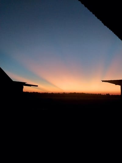 The image depicts a dusky sky with vibrant colors transitioning from a pale blue to a warm orange-pink glow near the horizon. This transition is illuminated by rays of light, suggesting a sunset or sunrise. In the foreground, the silhouette of a jagged, dark landmass, possibly a mountain range or a dense treeline, sharply divides the colorful sky from a completely black area on the right. The black area suggests a deep shadow, a very dark sky, or potentially an unlit portion of the scene. The sky takes up the majority of the image, with the dark silhouette forming a distinct vertical line across it. The overall mood is serene and perhaps a bit dramatic due to the stark contrast between the illuminated sky and the deep shadows. The time of day is clearly either dawn or dusk.