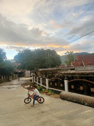This image depicts a tranquil residential street scene in Kecamatan Pangalengan, Indonesia, captured during the late afternoon or early evening. The sky is a prominent feature, filled with expansive clouds displaying a mix of grey, soft blue, and warm golden-orange tones, indicating either sunset or sunrise.
In the foreground, a young boy, wearing a white t-shirt, blue jeans, and sandals, is seen riding a red bicycle on a textured concrete road. He is positioned towards the left of the frame, facing right. Further down the street, another small figure on a bicycle can be faintly seen in the distance, heading away from the viewer.
To the right, there are houses with distinct reddish-brown tiled roofs. The property in the midground features a decorative black metal fence with ornate gold-colored patterns, topped by a simple roof extension. A large, grey pipe or culvert lies along the side of the road on the far right. The scene is rich with greenery, including dense trees that frame the houses and extend into the midground. Distant mountains or hills are visible in the background, softened by the atmospheric light. Overhead, power lines stretch across the sky. Near a junction or turn in the road, a white sign on a pole is visible, bearing unreadable text, likely in Indonesian. The scene portrays a sense of peaceful daily life in a semi-rural, hilly environment.
