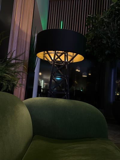 A modern interior scene with a large black drum-shaped lamp illuminating a wooden surface adorned with fern plants. The lamp has a golden interior visible through its intricate geometric support structure. In the background, a darkened cityscape is glimpsed, and a textured wall adds depth. To the right, a rounded, green upholstered chair provides a pop of color and softens the composition.