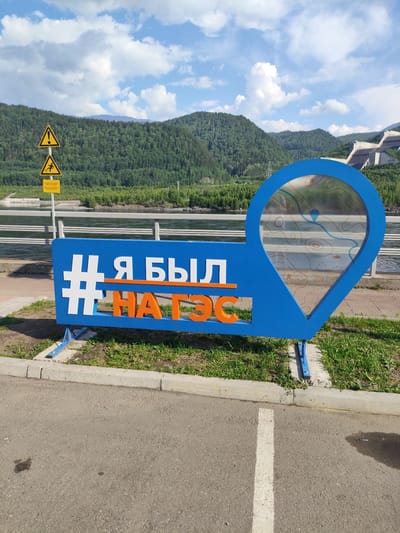 This media file is a photograph of a large, blue and orange sign with white lettering. The sign is shaped like a speech bubble with a map graphic inside a teardrop shape. The text on the sign appears to be in Cyrillic letters. The sign is placed outdoors on a sunny day with some clouds in the sky.
**Subjects:**
* **Objects:** The primary object is the prominent blue and orange sign. In the background, there are warning signs with yellow triangles, a metal railing, a body of water, a dam structure, and rolling, tree-covered hills.
* **Scene/Setting:** The scene is an outdoor area, likely a scenic viewpoint or park, with a body of water in the middle ground and mountains in the background. The ground in the foreground is paved asphalt, with a concrete curb and grass.
**Activity or Event:**
No specific activity or event is depicted, other than the sign being displayed. It appears to be a tourist attraction or informational sign.
**Notable Details:**
* **Time of Day:** The lighting suggests it is daytime, likely late morning or afternoon, given the brightness and shadows.
* **Weather:** The sky has blue patches and fluffy white clouds, indicating fair weather.
* **Location Cues:** The presence of a large dam structure, rolling forested hills, and the Cyrillic text on the sign suggest a location in Russia, possibly a hydroelectric power station. The provided location context of Cheremushki, Russia, is consistent with this. The map graphic within the sign might provide further local detail, showing a river and surrounding topography with a marker.
* **Emotions:** The overall impression is one of a pleasant, scenic outdoor location.
**Visible Text:**
The main sign displays the text: "# Я БЫЛ НА ГЭС". The warning signs in the background also have text, but it is too small to be clearly legible in the main image.