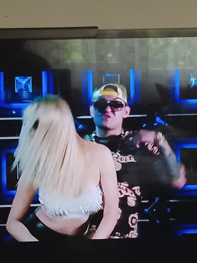 The image depicts a vibrant scene, likely from a music video or live performance, set in Karnalovo, Bulgaria. Two individuals are captured on what appears to be an illuminated stage or set.
In the foreground, a woman with long, flowing blonde hair is seen from behind, her head slightly turned. Her hair appears to be in motion, suggesting movement or dancing. She is wearing a strapless white crop top adorned with fringe or tassels, and black bottoms, possibly made of leather, revealing her lower back and midriff.
Behind her, to the right, is a man facing more towards the viewer. He wears a light-colored baseball cap backward, featuring a yellow detail on the rear. His eyes are obscured by dark sunglasses. He has visible tattoos covering his arms and is dressed in a dark t-shirt with a patterned design, along with a prominent chain or necklace around his neck. His right arm is raised and bent, making a gesturing motion with his hand.
The background features a dark space punctuated by vertical blue light bars and square or rectangular illuminated panels, creating a modern, dynamic, and artificially lit environment typical of an entertainment venue or set. The overall mood conveyed is energetic and performance-oriented. No discernible text is visible within the image.