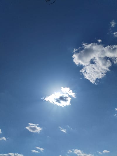 The image is an outdoor, vertical, low-angle shot of a bright blue sky with scattered clouds. The sun is partially obscured by a small, fluffy white cloud in the center of the frame, creating a lens flare and visible sunbeams. Several other clouds of varying sizes and densities are dispersed throughout the sky. The bottom portion of the image shows a sliver of a dark, silhouetted branch extending from the top left corner. The scene depicts a clear day with moderate cloud cover, suggesting a pleasant outdoor environment in Bilousivka, Ukraine. No people or specific objects other than the natural elements are visible.