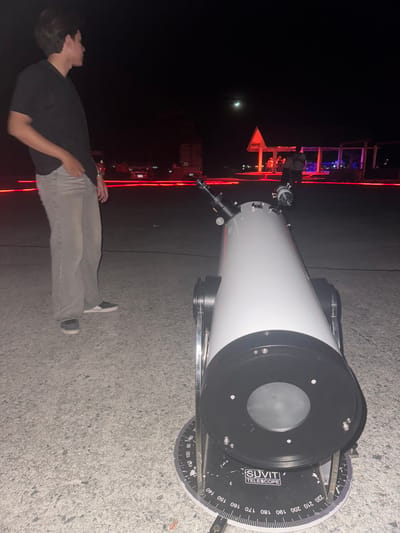 The image shows a person standing next to a telescope at night. The telescope is white with a black front and has a circular scale with markings that appear to be degrees. The person is wearing a black t-shirt and light-colored pants, looking up towards the sky. In the background, there are structures with red and blue lights, suggesting an outdoor event or area with illuminated features. The moon is visible in the dark sky. The ground appears to be asphalt or concrete. The overall scene suggests stargazing or an astronomical observation event. The visible text on the telescope reads "SUVIT TELESCOPE" and there are degree markings from 0 to 220. The time of day is clearly night. The location context provided is Baan Pang Ka, Thailand.