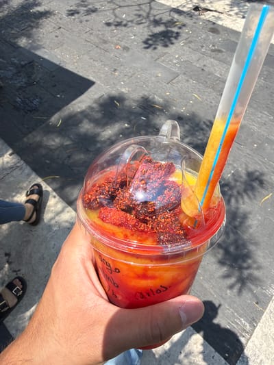 An outdoor, eye-level shot depicts a person holding a partially-consumed drink in Cuautla, Mexico. The drink, in a clear plastic cup with a dome lid, contains a yellow liquid at the bottom and a red liquid towards the top. The top portion of the drink is covered in chili powder. A clear plastic straw with a blue stripe runs through the drink, with the submerged portion tinted red and yellow. The cup has a name written on it.

The person's hand holds the cup; they are wearing a light blue denim material, likely jeans. Below the hand, one foot in a black sandal is visible on a grey paved surface. The environment includes a mix of sunlight and shadow.