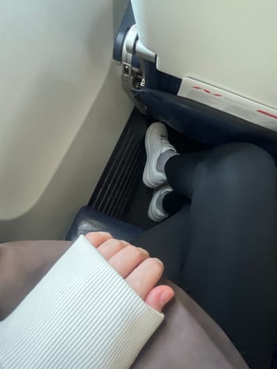The image shows a close-up view of a person's legs and feet, likely on a mode of transportation like an airplane or bus. The person is wearing dark leggings and white sneakers with gray socks. The setting appears to be indoors, with a white seat in the foreground and a dark blue seat in the background. A gray blanket or fabric is draped over the person's lap. The lighting suggests it might be daytime. A person's hand with manicured nails is visible on the right side of the frame, resting on the gray fabric. There is a folded piece of paper with red markings visible on the left side, possibly an in-flight safety card or informational pamphlet, with some text in an unreadable font. The overall impression is one of casual travel.