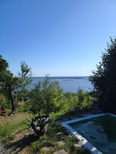 A wide-angle, eye-level shot captures a serene outdoor scene overlooking a body of water, likely the Dnieper River, near the village of Staiky, Ukraine. The sky is a clear, bright blue, dominating the upper portion of the frame. Lush green trees and foliage form a natural border along the foreground and mid-ground, with denser vegetation on the right side of the image.
In the foreground, to the left, a weathered tree stump with intricate, gnarled branches sits on a grassy slope. A wooden structure, possibly a hammock or a piece of garden furniture, is partially visible behind this stump. To the right, a small, rectangular swimming pool with a light blue border is filled with dark green water, reflecting the sky. The pool area is surrounded by stones and sparse vegetation.
The mid-ground features a slightly overgrown grassy area sloping down towards the expansive body of water. The water itself is calm and reflects the clear blue sky, with faint hints of landmasses or islands visible on the horizon. The overall impression is one of a peaceful, natural retreat. No people are visible in the image.