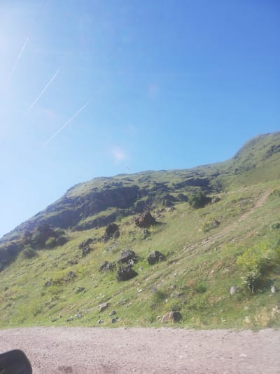 The image shows a natural landscape on a sunny day. A steep, grassy hillside with scattered rocks and boulders dominates the lower two-thirds of the frame. The hillside is covered in green vegetation with patches of yellow wildflowers. The top of the hill is obscured by shadow and the angle of the shot. In the sky, which is a clear, bright blue, there are several long, thin white contrails from airplanes. A single white bird is visible flying in the middle distance against the sky and hillside. In the foreground, a dirt or gravel road runs horizontally across the bottom of the image, with the edge of a black object, possibly a vehicle's side mirror, visible in the lower left corner. The overall impression is one of a rural or mountainous area. The location is Gogasheni, Georgia.
