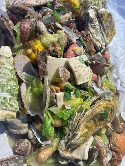 The scene presents a close-up, high-angle view of a large quantity of cooked seafood and mixed ingredients, located in Huangdao District, China. The food is spread across a wrinkled white sheet, possibly paper or plastic, which serves as a liner for an underlying surface. Dominant elements include numerous oysters, identified by their ridged shells, alongside various bivalve shells (clams or mussels) in differing states of openness. Also visible are cooked shrimp or prawns, and segments of a reddish-orange crab or crayfish. Interspersed among the seafood are multiple square-cut pieces of white tofu, some adorned with sesame seeds and chopped green herbs. Bright yellow corn kernels and substantial portions of cooked, light green leafy vegetables are also present. A viscous, pale green-yellow liquid or sauce uniformly coats the ingredients. The illumination is bright and direct, casting minimal shadows. No individuals or specific actions are depicted.