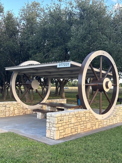 An exterior, eye-level, medium shot captures a picnic pavilion with a distinctive wagon wheel design, situated outdoors. The pavilion's roof is supported by large, decorative wagon wheels, one on each side, constructed from brown and tan material. These wheels are mounted on stone walls that form the base of the pavilion. Within the pavilion, a dark brown picnic table with two benches is visible, resting on concrete. A white sign with black numbers "077031" is attached to the underside of the pavilion's roof. The ground around the pavilion consists of concrete and green grass. In the background, numerous mature trees with dense foliage create a backdrop, and beyond them, a chain-link fence and a distant road are discernible under a clear blue sky with scattered clouds. A blue trash can is placed near the picnic table. The scene appears to be a public park or rest area. The location is identified as being in the city of Sonora, United States. There are no people visible in the image.