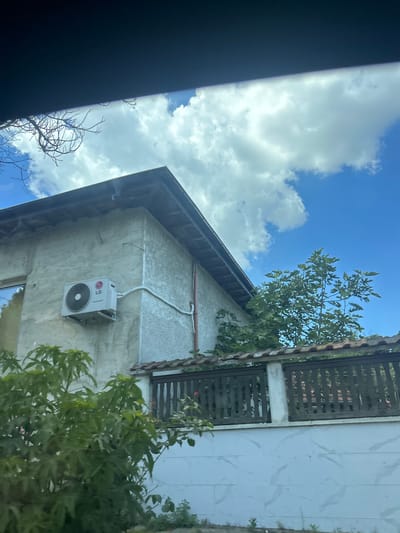 The image shows an exterior view of a building, likely a house, under a bright blue sky with scattered white clouds. The building appears to be constructed from concrete or stucco, with an aged, somewhat textured finish. An outdoor air conditioning unit, manufactured by LG, is mounted on the exterior wall. The roofline is visible, with dark shingles and visible wooden support beams. A fence with dark wooden slats runs horizontally across the mid-ground, partially obscuring a lower section of the building that is painted white with a marble-like pattern. Lush green foliage, including trees and bushes, surrounds the property, with some branches extending into the frame from the left and right. The overall impression is of a residential property in a sunny, possibly warm climate. The scene is likely located in the city of Beloslav, Bulgaria, based on the provided context, though no explicit landmarks are visible in the image to confirm this location. There are no people or animals visible in the frame.