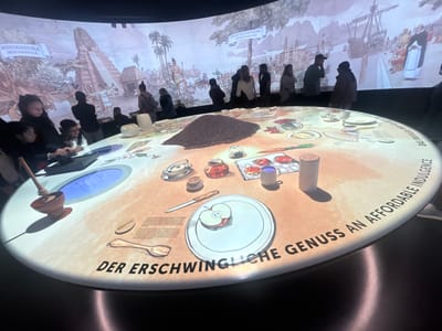 The image shows an exhibition or display, likely in a museum or cultural center, in Kilchberg (ZH), Switzerland. The display features a large, circular table with illustrations and text in German, including "Der erschwingliche Genuss an affordable indulgence." The tabletop features depictions of various foods and tools, including an apple on a plate, utensils, a mortar and pestle, and containers, all related to the history of chocolate. A pile of cocoa beans is visible near the center of the table.

In the background, a panoramic projection shows historical scenes related to Mesoamerica and Spanish expeditions, featuring images of pyramids, ships, and historical figures. Several people are standing around the table, observing the display and interacting with the exhibit. These individuals include adults and children. The scene is indoors, and the lighting is focused on the display table and projection screen, creating a bright and engaging environment.