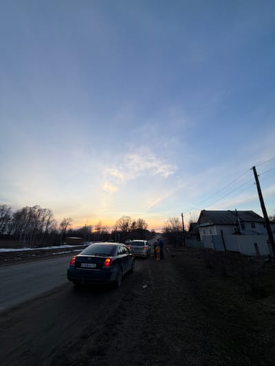 The image depicts a street in Дулесово, Russia, at dusk. The sky is a gradient of blue and orange, indicating a sunset. A grey, two-story building stands on the right side of the frame, adjacent to a grassy area. Utility poles with wires run alongside the building. Several cars are positioned on the road, with a dark-colored car being the closest to the viewer. The license plate on the back of this car shows "К 728 AP 10". Two people are standing next to one of the vehicles further down the road. Bare trees are visible in the background.