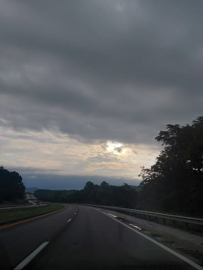 The image depicts a view from a vehicle traveling on a highway in Stanton, United States of America. The sky is overcast with dark clouds, but a patch of sunlight breaks through in the distance, illuminating the horizon. The highway curves to the left, with a metal guardrail running alongside the right shoulder. Puddles of water are visible on the shoulder, suggesting recent rain. The surrounding landscape is covered in dense green trees. A few vehicles, including a red semi-truck, are visible on the highway in the distance. The scene appears to be a rural or semi-rural setting, with the highway cutting through a forested area.