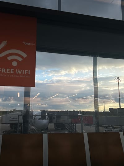 The image displays a view from inside an airport terminal, looking out through a large window onto an airport tarmac. The sky is partly cloudy, with visible patches of blue sky and sunlit clouds, suggesting it is daytime, possibly late afternoon or early evening.
An airplane, identifiable by its white nose cone and part of its fuselage, is parked at a gate. Adjacent to the aircraft is airport ground support equipment, specifically a catering or logistics truck that bears the text "VIE CATERING & LOGISTIC" on its side. In the distance, a red sign is partially visible, showing "V G" and "VIENNA". A tall pole with sensing equipment is also seen against the sky.
In the foreground, an orange sign with a white WiFi symbol is partially visible in the lower-left corner. The text on this sign clearly states "FREE WIFI" and "WIRELESS VIENNA AIRPORT", directly identifying the location as Vienna Airport. A logo resembling "VIE" is also present on the sign.
The scene captures an everyday airport operation at Vienna Airport in Fischamend, Austria, highlighting the availability of free Wi-Fi for passengers.