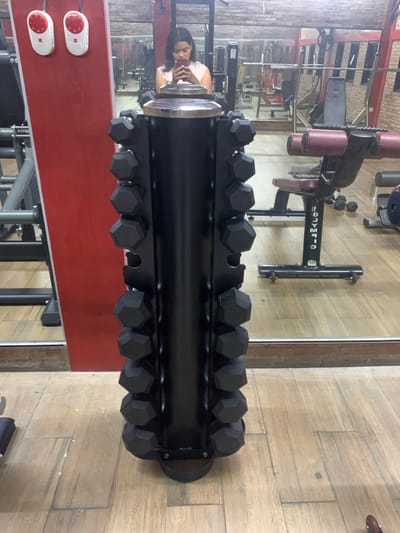The image shows a black dumbbell rack with black dumbbells arranged vertically in rows. The rack stands in a gym in Kecamatan Kartasura, Indonesia, at coordinates -7.567 latitude and 110.757 longitude. The rack is in front of a large mirror reflecting a woman in a pink shirt holding a smartphone. The gym has wooden floors and brick walls. An Olympic weight bench is in the background. The woman is reflected in the mirror. There is a red wall on the left with two white switches.