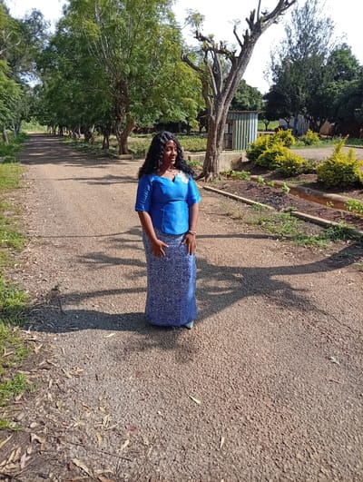 The image depicts a woman standing on a gravel path in what appears to be a semi-rural or park-like setting in Tambes Mile Four, Nigeria. She is wearing a vibrant blue top with a white embellishment on the shoulder and a long, patterned blue skirt. Her dark, curly hair frames her smiling face as she looks to her right. The woman is positioned in the center of the path, casting a distinct shadow on the ground.
The scene is characterized by lush green trees lining the path, creating a tunnel-like effect. Some of the trees are bare, showcasing their branching structures, while others are full of leaves. In the background, there are small structures, possibly sheds or buildings, and some low-lying yellow bushes. The ground is a mixture of gravel and dry leaves.
The lighting suggests it is daytime, likely late morning or early afternoon, given the angle of the shadows. The weather appears to be sunny and warm, contributing to the vibrant green of the foliage. The overall atmosphere is peaceful and serene. No visible text is present in the image.