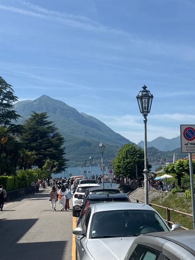 Captured from a street level perspective in the Italian town of Gittana, the image presents a landscape dominated by mountains, buildings, and a row of parked vehicles. A line of people is walking down the street to the left of the parked vehicles. The backdrop features a mountain range under a clear blue sky. Closer to the foreground, trees and a streetlamp with a decorative design are seen. A "No Parking" sign with a red circle and diagonal line is visible above the street lamp. The photo's orientation is vertical.