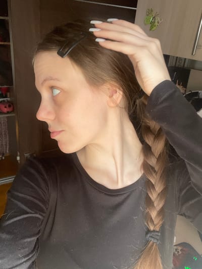 This image, rotated 90 degrees clockwise, presents a side profile of a young person, likely female, in an indoor setting in Ulukulevo, Russia. The subject has long, light brown hair styled in a single, thick braid, secured at the end with a black hair tie. A black hair clip is visible near the forehead, holding back some hair.

The person is wearing a plain black long-sleeved top. One hand, with long, light-colored (white or light grey) nail polish, is raised and positioned near the head, seemingly adjusting the hair or posing. The subject has fair skin and a soft profile, with closed lips and the eye partially visible, looking downwards.

The background consists of a warm-toned, possibly wooden or laminate surface, likely part of a furniture piece such as a wardrobe or shelf. A small, decorative green butterfly is affixed to this surface. The lighting suggests an indoor environment, without specific cues for time of day or weather. There is no visible text in the image.