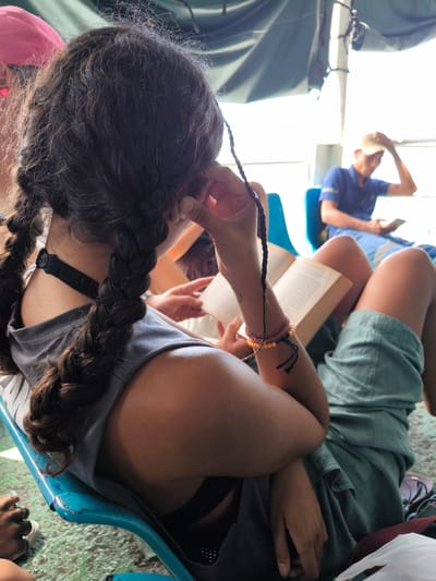 A woman with dark hair in braids sits on a blue plastic chair, reading a book. She wears a grey shirt, green shorts, and a black choker. A man sits in a blue chair in the background, wearing a blue shirt and khaki hat, looking at a phone or book. The setting appears to be indoors with bright natural light. The location is Gunung Malang, Indonesia.
