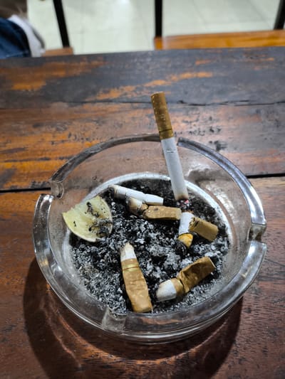 The image shows a glass ashtray on a weathered, dark wooden table. The ashtray is filled with cigarette butts, ash, and a small piece of lemon rind. One cigarette is still burning upright within the ashtray. The setting is unclear, and there is no verifiable connection to the city of Perugia, Italy within the visible context.