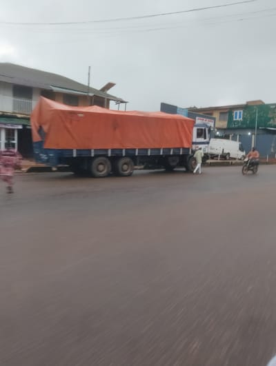 The image depicts a street scene in Aketu-Oja, Nigeria. A large truck with an orange tarp covering its cargo is the central focus. The truck has multiple wheels and is positioned on a wet road, suggesting recent rain. To the left of the truck, a blurred figure, possibly a person, is visible. A man in a light-colored shirt and pants is standing near the truck's front tire. Further to the right, a person on a motorcycle is riding away from the camera. In the background, there are buildings, including one with a blue window and another with a green facade. The sky is overcast. The road surface is dark and appears wet. The overall impression is of a busy, possibly commercial area in Nigeria.