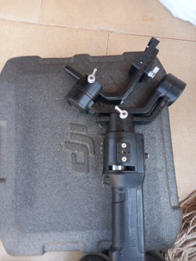 The image shows a black DJI Ronin-M camera stabilizer, lying on its side inside a black foam-lined carrying case. The stabilizer is made of black plastic and metal, with a silver knob on the left side and two silver knobs on the right side. The stabilizer is labeled “RONIN-M”. The case is made of black plastic and has a textured foam lining. The image is likely taken in Ita Oniyan, Nigeria, as the provided coordinates are located within the city. The exact location cannot be determined from the image alone. There are no people or other objects visible in the image, other than the stabilizer and its case.