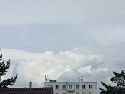 The image depicts an outdoor scene with a cloudy sky dominating the upper portion. Below the sky, the roofline of a building is visible, along with parts of a pine tree on the left and right edges of the frame. The building appears to be a multi-story structure with a brick facade. Several antennas and communication equipment are mounted on its roof. The foreground features a rusty, corrugated metal roof. The scene is devoid of any people or explicit actions. The lighting suggests it is daytime, possibly overcast or approaching dusk. The setting appears to be a residential area, potentially in the city of Kenjekol, Kazakhstan, given the context provided.