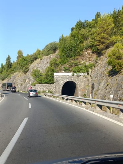 The image captures a scenic view of a road leading to a tunnel carved into a rocky hillside. The road is paved and has white lane markings. A gray car is driving in the right lane, and a black bus is in the left lane. The hillside is covered with lush green trees and vegetation. The tunnel entrance is arched and made of stone. A guardrail runs alongside the road. The sky is clear and blue. The scene appears to be located in Kruševica, Montenegro, given the context of the prompt. The overall impression is one of a peaceful, natural environment with a road and tunnel providing passage through the landscape.