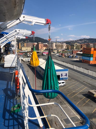 The image captures a daytime scene at the Port of La Spezia, Italy, observed from the deck of a large vessel. In the foreground, two distinct davits, white with red accents, extend from the ship's port side. From these davits, two large, teardrop-shaped fabric bags—one yellow and one teal-green—are suspended by chains and shackles, hanging over the ship's railing towards the dock. A partial view of a white lifeboat is visible above the davits. On the blue deck to the left, an orange life buoy is secured to the white railing, and a green bucket rests on the deck. Three individuals are visible standing on the ship's deck in the upper-left quadrant, dressed in dark clothing, appearing to observe the operation.
Beyond the ship, a paved port area stretches under a clear blue sky with scattered white clouds. This area includes a road where several vehicles, including white vans and a flatbed truck loaded with materials, are present. A low-rise building with a blue facade displaying the white text "SPEZIA ZAMBRA" is prominent in the mid-ground. To the right, multiple stacks of intermodal shipping containers are visible, bearing logos such as Hapag-Lloyd, UASC, and Maersk. The background reveals the city of La Spezia, characterized by multi-story buildings with varied facades, some featuring reddish-brown roofs, which ascend into verdant hills. The scene is brightly illuminated by sunlight, casting sharp shadows from the ship's structures and other objects.
