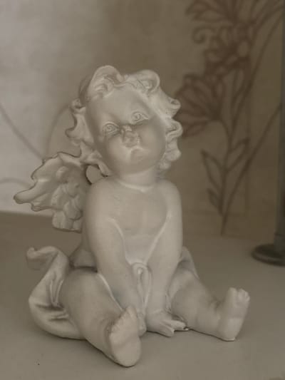 The image displays a close-up, eye-level, indoor shot of a white, plaster cherub figurine. The figurine is depicted in a seated position, with its legs crossed and its arms resting on its knees. It has small, detailed wings folded against its back and curly hair. The background is softly lit and shows a blurred floral pattern on what appears to be wallpaper. The surface on which the figurine rests is a pale, flat surface. The image quality is slightly grainy. No people, actions, or interactions are visible, and the location of Tikhiye Zori, Russia, is not depicted or suggested by the visual information within the image.