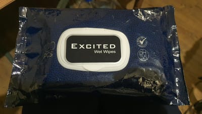 The image shows a package of "EXCITED Wet Wipes" with 120 pieces. The package is dark blue with a pattern of small black dots. A white rectangular tab is on the front of the package, revealing the opening for the wipes. To the left of the tab, "120 PCS" is written in white text. To the right of the tab, there are two circular logos: one with a checkmark and text that appears to be "ALCOHOL FREE," and another with "pH 5.5" written inside a circle. The package is slightly creased and wrinkled, suggesting it is a flexible plastic material. The background is out of focus and appears to be a wooden surface with some dark areas. There is no indication of the location being in the city of Karankul, Uzbekistan.