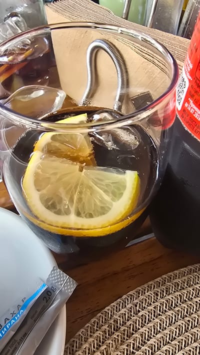 In the city of Asparuhovo, Bulgaria, on a wooden table surface, a close-up shot focuses on a clear glass containing a beverage. The drink is filled with ice cubes and garnished with lemon slices, one of which is prominently featured in the foreground, showcasing its vibrant yellow rind and translucent flesh. The glass is partially submerged in what appears to be a dark liquid. Behind the glass, a metal spring-like object emerges from a stack of light brown paper napkins. To the right of the glass, a portion of a red-labeled bottle is visible, its texture appearing slightly wet. In the lower left corner, a white plate holds two small, silver packets, one with blue lettering and the other with gray lettering, one of which indicates "2,0g." A woven placemat occupies the lower right portion of the frame. The background is softly blurred, suggesting an indoor setting with ambient lighting.