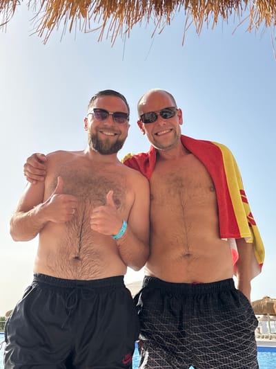 Two men, shirtless and wearing sunglasses, pose for a photo outdoors in Al Ahia', Egypt. The top man is bald and has a red and yellow towel draped over his shoulders. The bottom man has a beard. Both men are wearing black swim trunks. The bottom man gives two thumbs up. The sky is clear and bright. A thatched roof structure is visible in the upper left corner. A building and swimming pool are visible in the upper right corner.