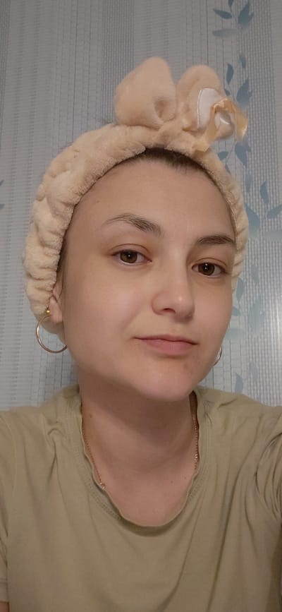 A young woman is pictured from the chest up, looking slightly to her right. She is wearing a beige fuzzy headband with a bow detail on top, and a light green t-shirt with a thin gold chain necklace. She has brown eyes and light skin, and is wearing hoop earrings. The background is a blue, textured wallpaper with a subtle floral pattern. The lighting is soft and appears to be artificial. The image is a close-up portrait, focusing on the woman's face and upper body.
