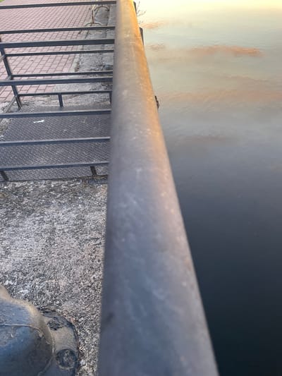 The image shows a metal railing and stairs overlooking a body of water. The stairs are made of metal grating and diamond plate, with a paved area beyond them. The water reflects the sky, with clouds visible. There is a dark, round object at the bottom left of the image, which appears to be part of a railing support or bollard. The scene is outdoors, likely a waterfront area. The location is the city of Sovetsk, Russia.