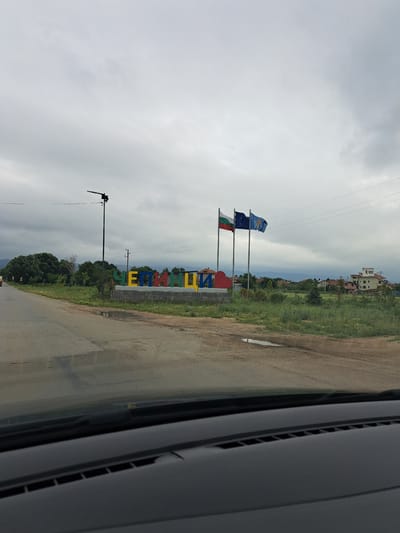 The image captures a roadside view in Dolni Bogrov, Bulgaria, on a cloudy day. A large, colorful sign spelling out "ЧЕПИНЦИ" (Chepinsi) in Cyrillic letters stands prominently on the right side of the road, constructed from individual, brightly colored blocks (yellow, blue, red, yellow, blue, red, yellow). Adjacent to the sign, three flags are flown on tall poles: the Bulgarian flag (white, green, red), the European Union flag, and another flag, possibly a regional or municipal one, in blue with a yellow emblem.
In the background, the scene features residential buildings with reddish-tiled roofs, a cluster of green trees, and a distant view of hills or mountains under the overcast sky. The foreground shows a paved road with some cracks and puddles, suggesting recent rain. A portion of a car's dashboard and windshield is visible at the very bottom of the image, indicating the photo was taken from inside a vehicle. A street lamp is positioned behind the sign, and utility poles with wires run across the scene. The overall environment appears to be a semi-rural or suburban area on the outskirts of a town.